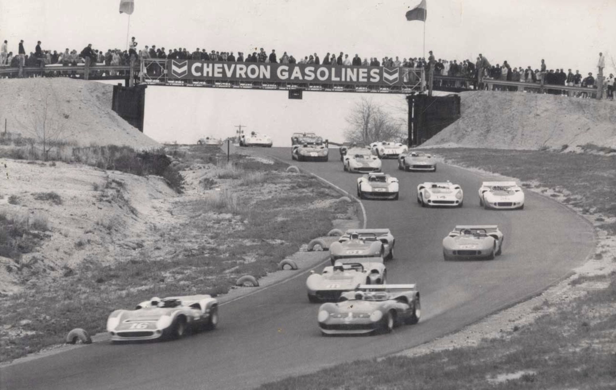 Black-and-white photo of sports racing cars passing under the Chevron Gasolines bridge at the Bridgehampton race circuit, crowds watching from above